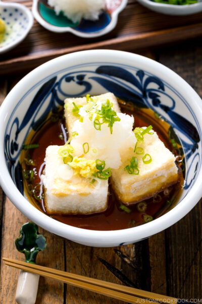 A ceramic bowl containing Agedashi Tofu (Agedashi Dofu) topped with grated daikon, chopped scallions, and grated ginger.