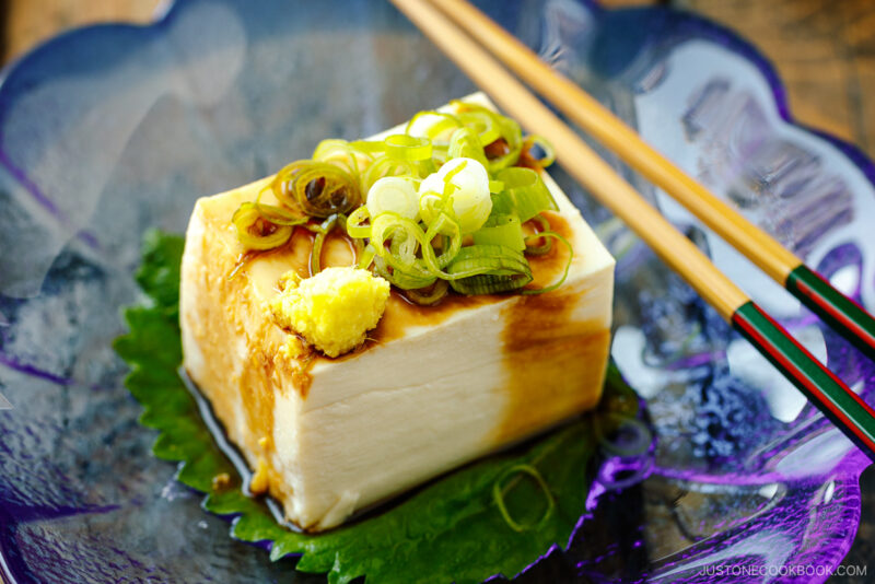 A glassware containing Japanese cold tofu topped with green onion and grated ginger and drizzled with soy sauce.