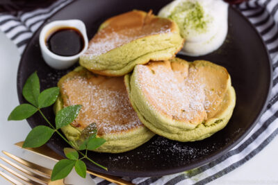 A black plate containing 3 matcha souffle pancakes with fresh whipped cream and maple syrup on the side.