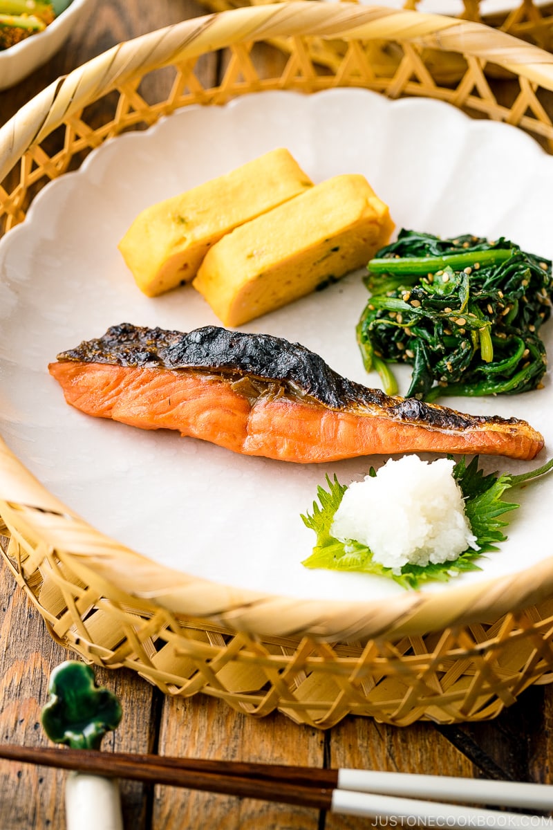 A white plate containing Japanese salted salmon (Shiozake), tamagoyaki, and spinach salad.