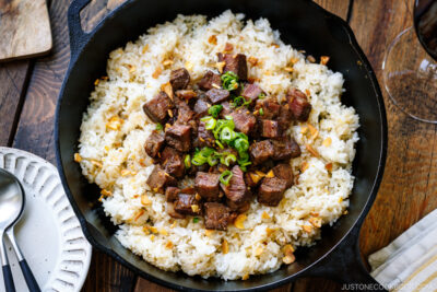 A cast-iron pan containing Steak Garlic Rice topped with chopped scallions and garlic chips.