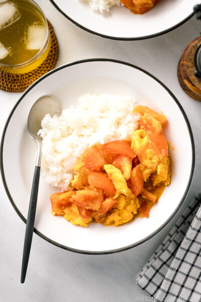 A white ceramic shallow bowl containing Stir-Fried Tomatoes and Eggs over steamed rice.