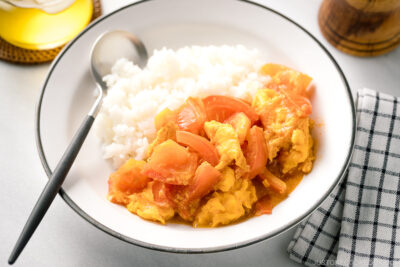 A white ceramic shallow bowl containing Stir-Fried Tomatoes and Eggs over steamed rice.