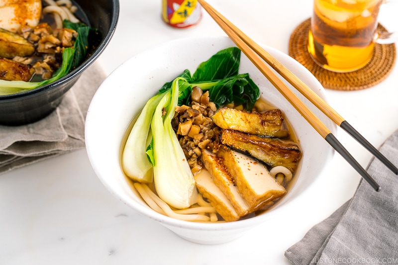 A white bowl containing Vegetarian Udon Noodle Soup topped with bok choy, fried tofu, minced mushroom, and crispy eggplant.