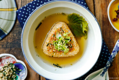 A donburi bowl containing a Japanese grilled rice ball in a flavorful dashi broth, topped with green onions and furikake rice seasoning.