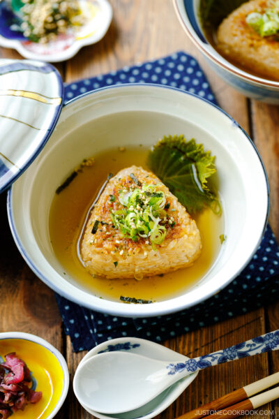 A donburi bowl containing a Japanese grilled rice ball in a flavorful dashi broth, topped with green onions and furikake rice seasoning.